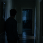 A person standing alone in a dimly lit hallway after hearing their name called from an empty room.